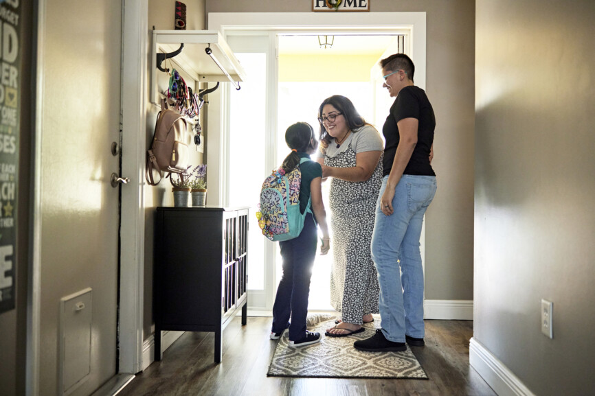 Full length view of smiling women standing at sunny entrance to family home and helping 8 year old girl with backpack before leaving.