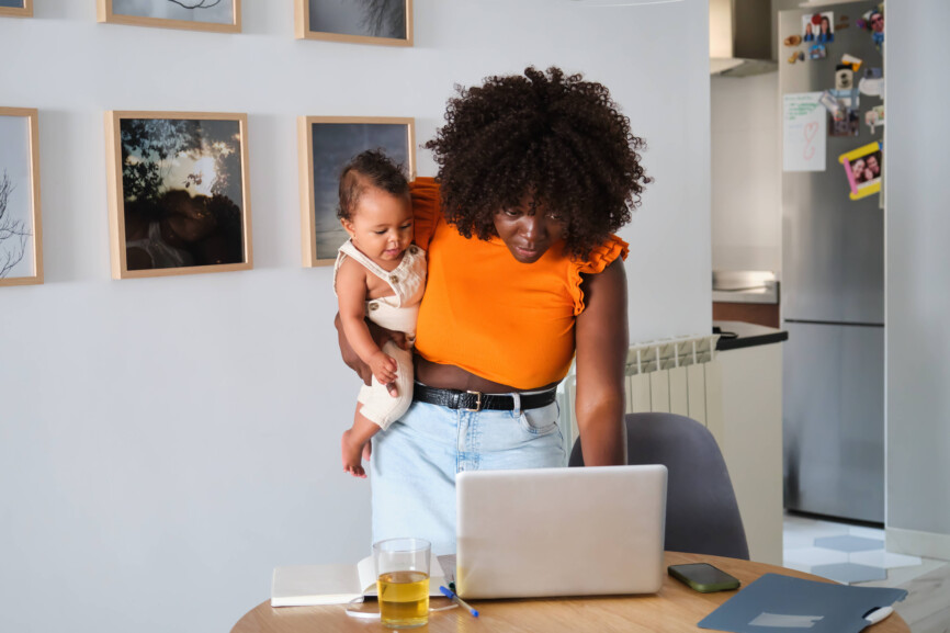 An African mother multitasks, working on her laptop while carrying her baby girl in a modern home setting, showing a blend of work and family life Model Released HaruUraraStudio_ID24141_699400_025 Copyright: xHaruxUraraxStudiox RECORD DATE NOT STATED